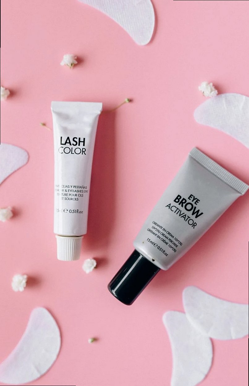 Plano cenital de tubos cosméticos para pestañas y cejas sobre fondo rosa con detalles florales.